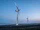 Low-angle view of a four-blade wind turbine in a wind farm at dusk under a blue-gray sky.