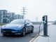 Electric vehicle charging at a roadside pedestal with nearby electrical substation and overhead power lines suggesting the grid infrastructure behind charging capacity.