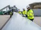 Workers inspecting a decommissioned wind turbine blade segment in a recycling yard with recycling equipment in the background.